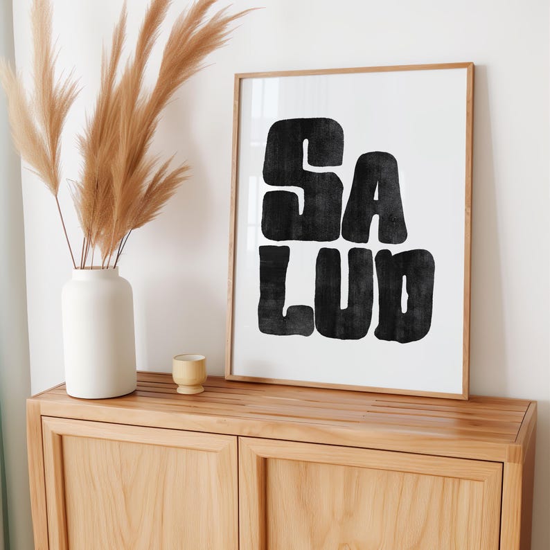 Op de afbeelding: Een ingelijst zwart-witdruk met het woord "SALUD" in vetgedrukte blokletters. De print hangt aan een licht houten muur boven een licht houten kast.