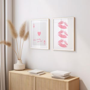 May include: Two framed art prints on a wall. One features a playing card with a heart and the text "How lucky are we?" The other print displays three pink lip prints. The frames have a gold finish.