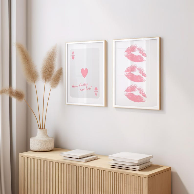 May include: Two framed art prints on a wall. One features a playing card with a heart and the text "How lucky are we?" The other print displays three pink lip prints. The frames have a gold finish.