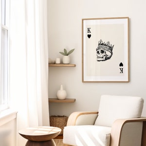 May include: A black and white illustration of a skull wearing a crown, with the text "K" and a spade symbol on the card. The card is framed in a light wood frame and hanging on a white wall.