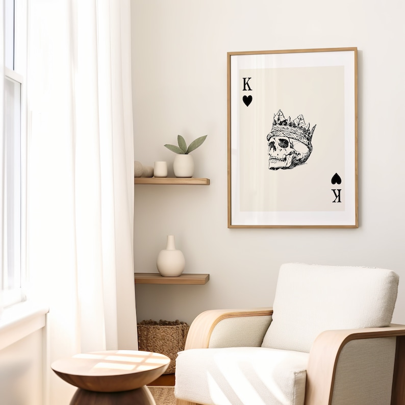 May include: A black and white illustration of a skull wearing a crown, with the text "K" and a spade symbol on the card. The card is framed in a light wood frame and hanging on a white wall.