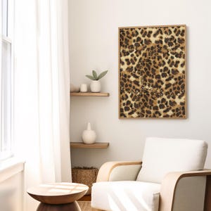 May include: A framed print of a leopard print pattern in shades of brown, tan, and white. The print is hanging on a white wall in a room with a white armchair and a wooden side table.