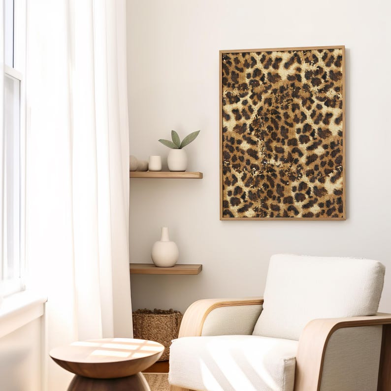 May include: A framed print of a leopard print pattern in shades of brown, tan, and white. The print is hanging on a white wall in a room with a white armchair and a wooden side table.