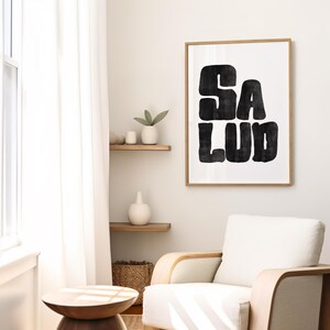 Op de afbeelding: Een zwart-witte kunstprint met het woord "SALUD" in een vet, gestileerd lettertype. De print is ingelijst in een licht houten lijst en hangt aan een witte muur.