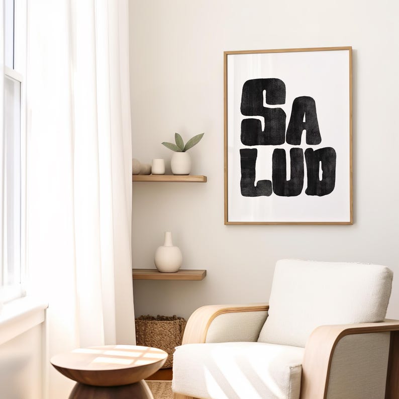 Op de afbeelding: Een zwart-witte kunstprint met het woord "SALUD" in een vet, gestileerd lettertype. De print is ingelijst in een licht houten lijst en hangt aan een witte muur.