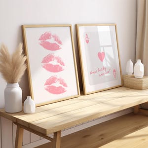 May include: Two framed art prints. One features three pink lip prints on a white background. The other displays an Ace of Hearts card with the text "How lucky are we?" in pink. Both are in light wood frames.