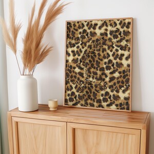 May include: A framed print featuring a brown and gold leopard print pattern. The print is hanging on a light wood cabinet with a vase of dried pampas grass.