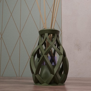 Puede incluir: Un difusor de caña verde oliva con un diseño decorativo calado. El recipiente de vidrio contiene cañas y un líquido púrpura. El difusor está sobre una superficie de madera, con una pared estampada en el fondo.