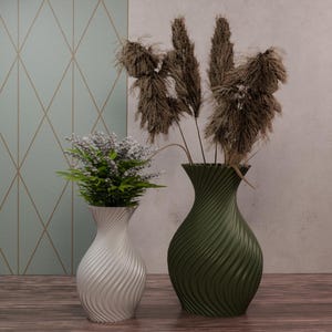 Peut inclure: Deux vases décoratifs, un blanc et un vert olive, sont posés sur une surface en bois. Le vase blanc contient une petite plante avec des feuilles vertes et des fleurs violettes. Le vase vert contient de hautes herbes brunes séchées. Les vases ont un design tourbillonnant texturé.
