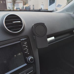 Puede incluir: Soporte negro para teléfono de coche, fijado al salpicadero. El soporte tiene una zona magnética circular y una base rectangular. El interior del coche incluye una pantalla de navegación, una rejilla de ventilación y botones.