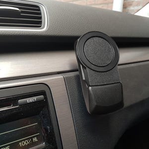 Peut inclure: Un support de téléphone noir pour voiture fixé sur le tableau de bord. Le support a une zone magnétique circulaire et une base rectangulaire. Le tableau de bord est gris foncé avec une garniture argentée et une bouche d'aération.