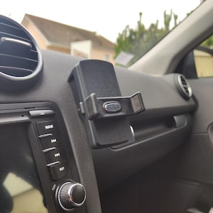 May include: A black car phone holder mounted on a car dashboard. The holder is attached to the dashboard with a clip and has adjustable arms to secure a phone. The dashboard also features a stereo system and air vents.