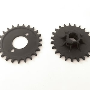 Peut inclure: Deux pignons circulaires noirs avec des dents d'engrenage sur le bord extérieur. Le pignon de gauche présente une face plate avec un trou central et deux petites ouvertures circulaires. Celui de droite montre un moyeu central surélevé avec des ailettes.