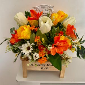 May include: A floral arrangement in a wooden planter. The arrangement features orange, yellow, and white flowers, with green foliage. The planter has the words "In loving memory Dad" and a decorative sign that says "Special Dad".
