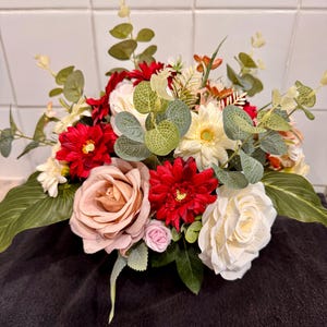 May include: A colourful floral arrangement with red dahlias, cream roses, and blush roses. Green leaves and eucalyptus sprigs add texture. The bouquet is set against a dark background, highlighting the colourful blooms.