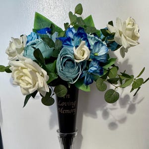 May include: A floral arrangement featuring white, teal, and blue roses, hydrangeas, and greenery. The flowers are arranged in a black vase with the words "In Loving Memory" printed on it. The arrangement is set against a white background.