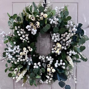 Peut inclure: Une couronne décorative circulaire composée d'un mélange de verdure et de baies blanches. La couronne comprend des feuilles d'eucalyptus vertes, des aiguilles de pin et des baies blanches et crème. Accrochée à une porte blanche.