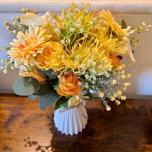 Può includere: Un vivace bouquet di fiori gialli e arancioni, tra cui rose e margherite, disposto in un vaso bianco strutturato. Il vaso è legato con spago. I fiori sono appoggiati su una superficie di legno scuro.
