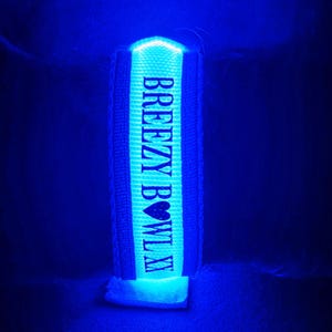 Può includere: Un braccialetto blu luminoso con la scritta "BREEZY BOWLAX" in bianco. Il braccialetto è centrato nell'immagine, con le parole "CUSTOM TEXT" sopra e sotto. Lo sfondo è blu scuro.
