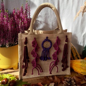 May include: A beige jute tote bag with a woven handle, decorated with purple and burgundy yarn tassels and a dreamcatcher design. The bag is set against a backdrop of fall foliage, pumpkins, and a pot of purple heather.