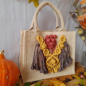 Può includere: Una borsa tote in iuta beige con un design macramè giallo, bordeaux e marrone. La borsa ha una forma rettangolare con un manico. Una zucca arancione e il fogliame autunnale sullo sfondo.
