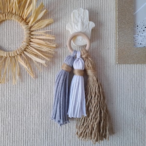 Może przedstawiać: Biała ceramiczna ręka Hamsa z drewnianym pierścieniem wiszącym na niej. Trzy frędzle wiszą z pierścienia, jeden jest biały, jeden jest jasnoszary, a jeden jest brązowy.