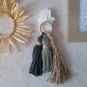 Può includere: Un gancio da parete in ceramica bianca a forma di mano con tre nappe appese. Le nappe sono verdi, verde oliva e beige.