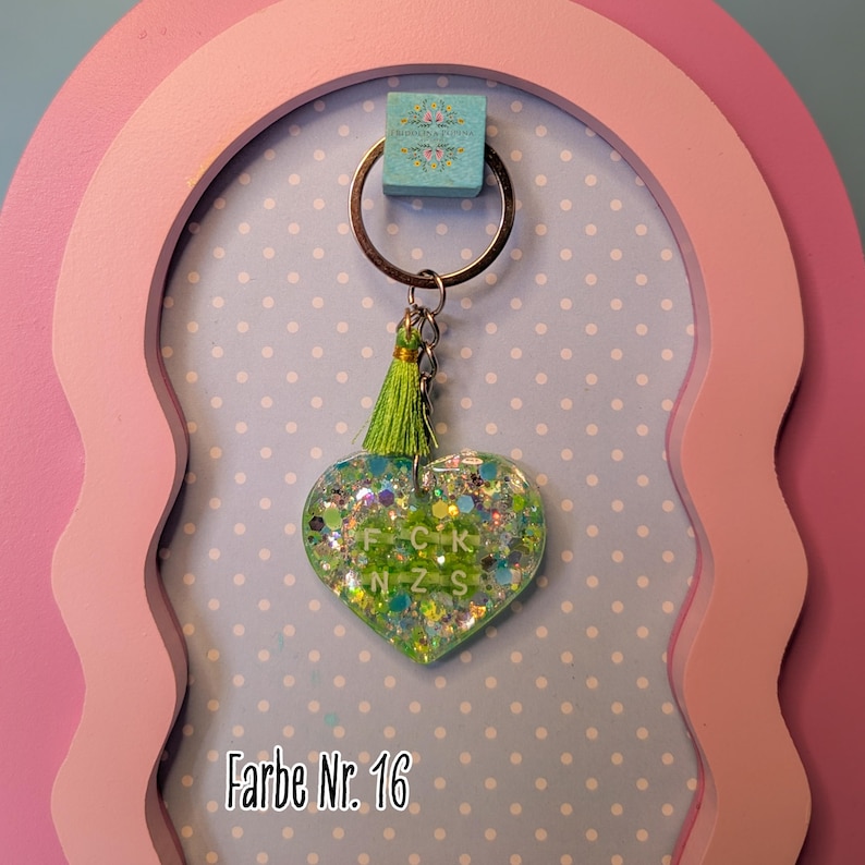 Op de afbeelding: Een hartvormige sleutelhanger van heldere hars gevuld met groene en blauwe glitters, voorzien van een limoengroene kwast en een zilverkleurige metalen ring. Het hart bevat witte letterkralen die FCK NZS spellen. Tekst Farbe Nr. 16 is zichtbaar.