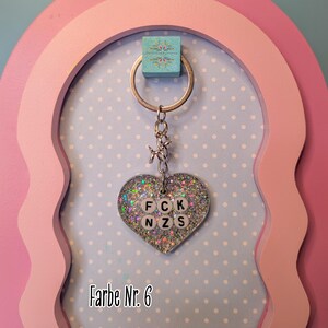 Op de afbeelding: Een hartvormige sleutelhanger met zilveren glitters in transparante hars. In het midden zitten witte ronde kralen met zwarte letters die FCK NZS spellen. De hanger hangt aan een zilverkleurige metalen ring met een kleine verbinding.