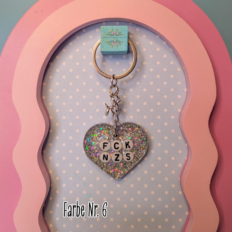 Op de afbeelding: Een hartvormige sleutelhanger met zilveren glitters in transparante hars. In het midden zitten witte ronde kralen met zwarte letters die FCK NZS spellen. De hanger hangt aan een zilverkleurige metalen ring met een kleine verbinding.