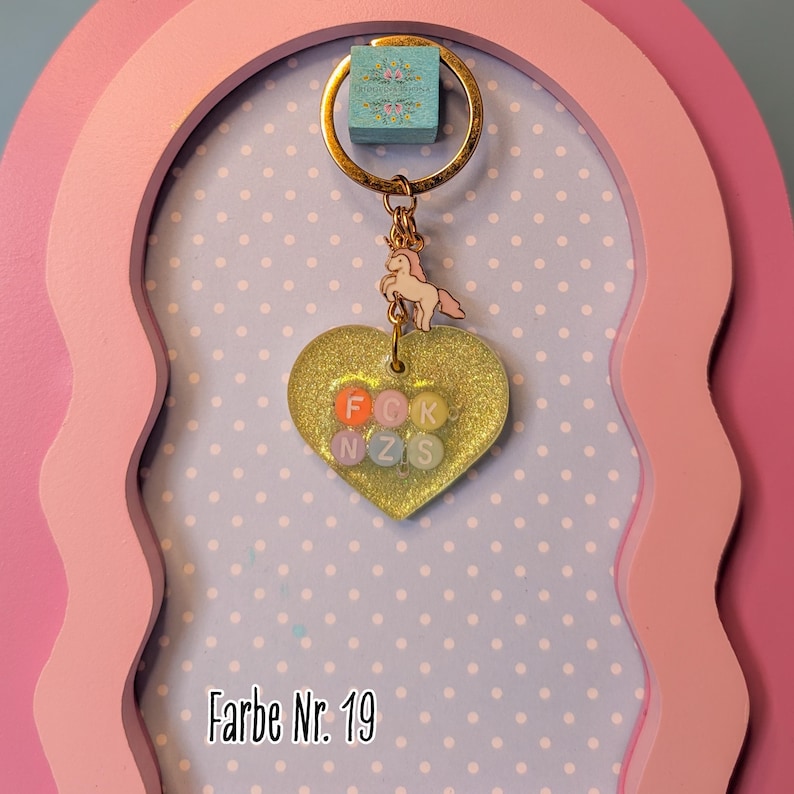 Op de afbeelding: Een goudkleurige metalen sleutelhanger met een kleine roze paardenbedel en een hartvormige hanger. Het hart is gemaakt van doorschijnende gele glitterhars en bevat zes kleine ronde kralen met de letters F, C, K, N, Z en S erop gedrukt.
