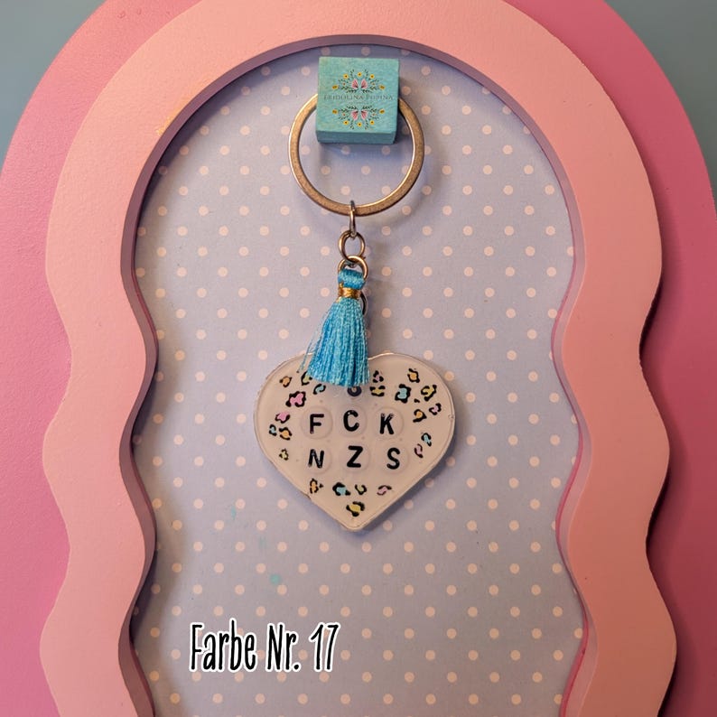 Op de afbeelding: Een hartvormige sleutelhanger met een witte achtergrond en een kleurrijk luipaardpatroon. In het midden staan zwarte letterkralen met de tekst F C K N Z S. Een lichtblauw kwastje hangt aan de zilverkleurige metalen ring, bevestigd aan een klein vierkant blauw label.