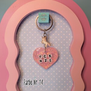 Op de afbeelding: Een hartvormige sleutelhanger van roze glitterhars met een kleine metalen eenhoornbedel aan een zilveren ring. Het hars hart bevat witte letterkralen in twee rijen die FCK NZS spellen. De tekst Farbe Nr. 14 is zichtbaar op de afbeelding.