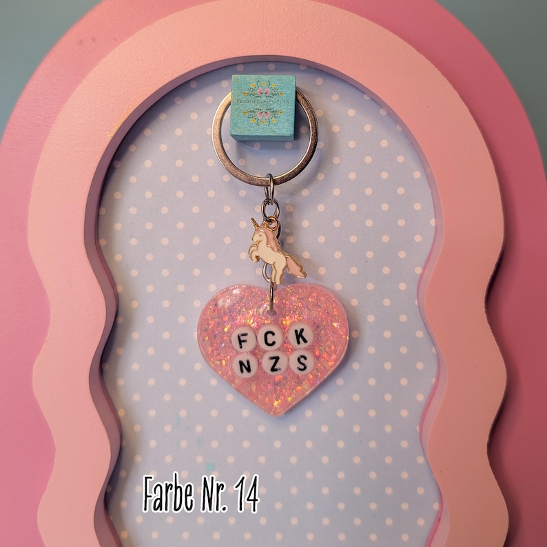 Op de afbeelding: Een hartvormige sleutelhanger van roze glitterhars met een kleine metalen eenhoornbedel aan een zilveren ring. Het hars hart bevat witte letterkralen in twee rijen die FCK NZS spellen. De tekst Farbe Nr. 14 is zichtbaar op de afbeelding.