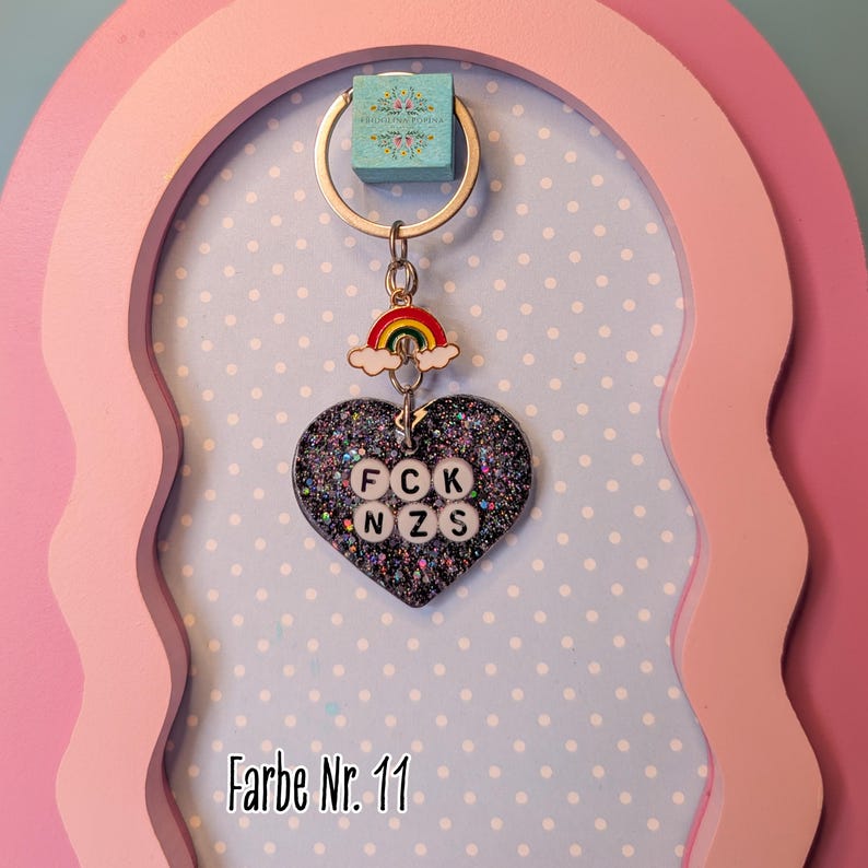 Op de afbeelding: Een hartvormige sleutelhanger met een zwarte glitterafwerking en ingebedde witte ronde kralen met de letters FCK NZS. De sleutelhanger bevat een kleine metalen regenboogbedel bevestigd aan een zilverkleurige ring en een blauw label.
