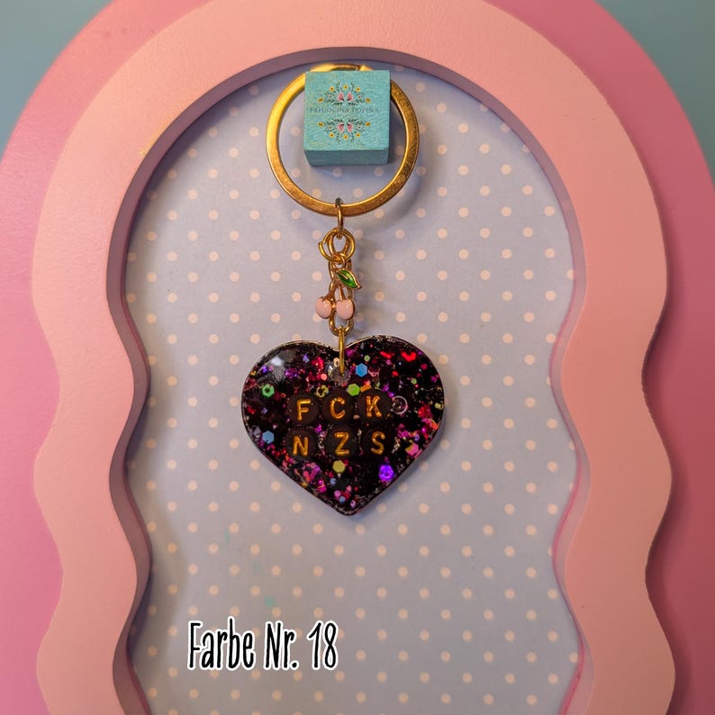 Op de afbeelding: Een hartvormige sleutelhanger van zwarte hars gevuld met veelkleurige glitters en kleine zwarte kralen. De kralen bevatten de letters F, C, K, N, Z en S in het oranje. Een goudkleurige metalen ring en een kleine kersenbedel zijn aan de bovenkant bevestigd.