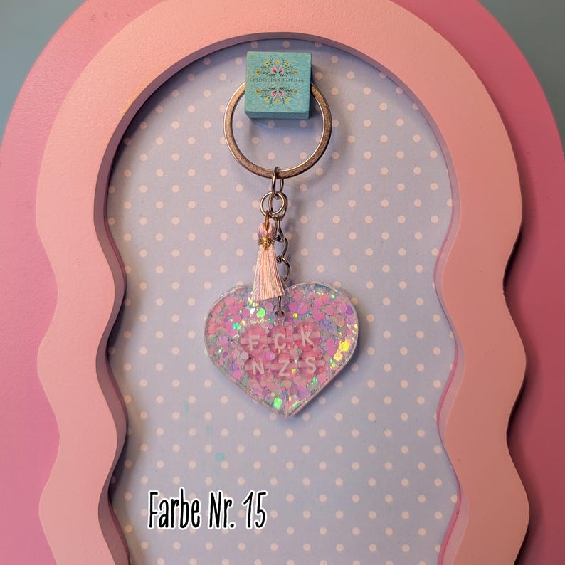 Op de afbeelding: Een hartvormige sleutelhanger met een doorzichtige harsbehuizing gevuld met iriserende roze en paarse glitters. In het midden zitten witte letterkralen die FCK NZS spellen. Een klein, lichtroze kwastje hangt aan de zilverkleurige metalen ring.