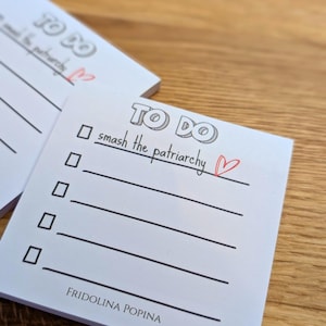 Può includere: Blocco note bianco con le parole "TO DO" in un font a bolle. Il primo elemento dell'elenco è "smash the patriarchy" con un cuore disegnato accanto. Il blocco note è su una superficie di legno.