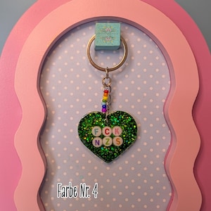 Op de afbeelding: Een hartvormige sleutelhanger met een donkergroene glitterafwerking. In het midden bevinden zich witte ronde kralen met de letters F, C, K, N, Z en S in verschillende kleuren. Een klein kettinkje met regenboogkleurige kralen verbindt het hart met een metalen ring.