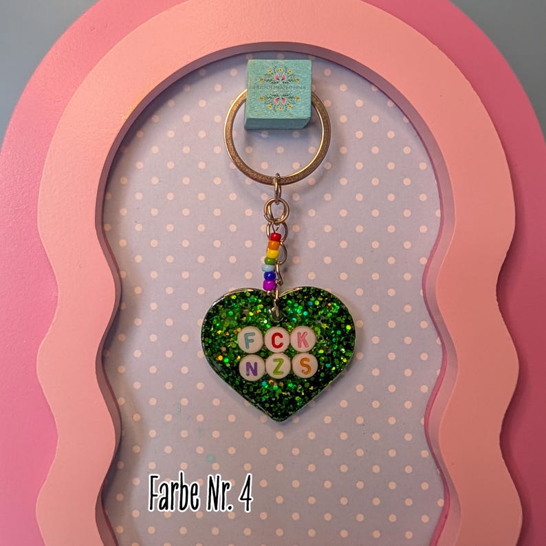 Op de afbeelding: Een hartvormige sleutelhanger met een donkergroene glitterafwerking. In het midden bevinden zich witte ronde kralen met de letters F, C, K, N, Z en S in verschillende kleuren. Een klein kettinkje met regenboogkleurige kralen verbindt het hart met een metalen ring.
