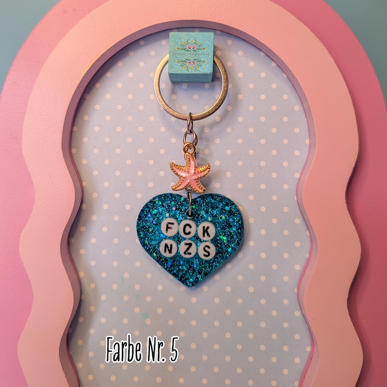 Op de afbeelding: Een hartvormige sleutelhanger van blauwe glitterhars met een kleine roze zeesterbedel en witte letterkralen die FCK NZS spellen. De sleutelhanger is bevestigd aan een zilverkleurige metalen ring en hangt tegen een lichtblauwe achtergrond met stippen.