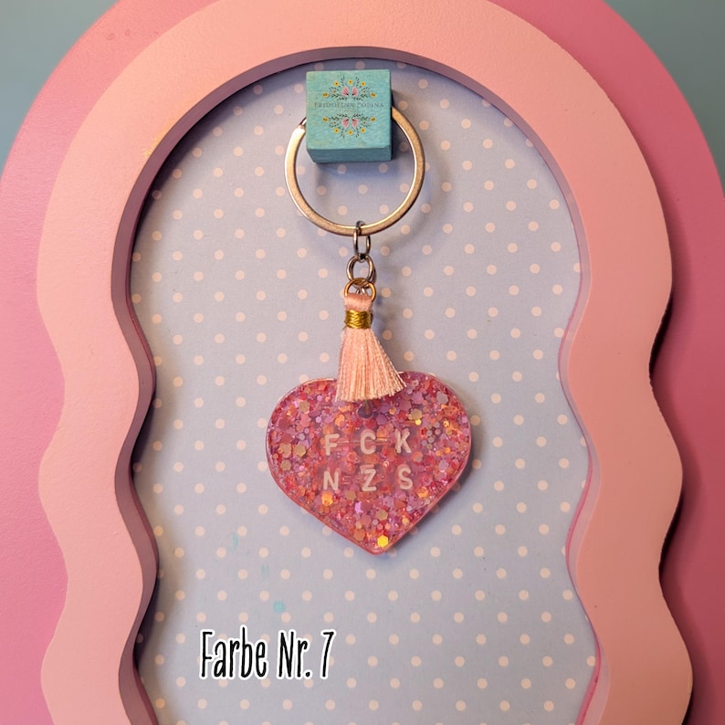 Op de afbeelding: Een hartvormige sleutelhanger van roze hars gevuld met iriserende glitters. De sleutelhanger bevat de witte letters F C K en N Z S in het midden, een klein lichtroze kwastje met gouddraad en een zilveren metalen ring.