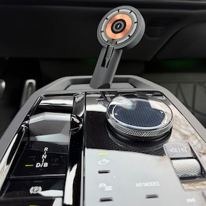 May include: A grey and copper wireless phone charger mounted in a car's console. The charger has a circular design with a copper centre and a grey stem. The car's console features a media control knob and gear shift controls.