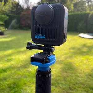 May include: A black GoPro Max action camera with a circular lens cover, mounted on a black and blue extension pole. The camera features a vertical ribbed design on the front, the GoPro logo in blue, and a side button with the text MAX printed in blue.