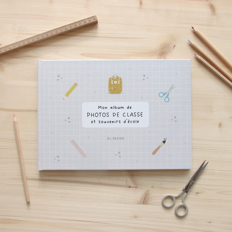 Puede incluir: Un &aacute;lbum de fotos gris con un patr&oacute;n de cuadr&iacute;cula y un t&iacute;tulo que dice "Mon album de PHOTOS DE CLASSE et souvenirs d'&eacute;cole" con ilustraciones de mochila, l&aacute;piz, tijeras y pincel. El &aacute;lbum est&aacute; sobre una superficie de madera clara.