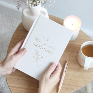 Peut inclure: Un journal blanc avec l'inscription dorée "Mon journal de GRATITUDE" et une illustration dorée d'une main tenant une feuille et des étoiles.