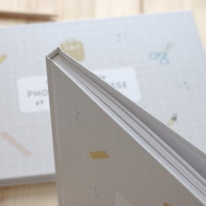 Puede incluir: Primer plano de un &aacute;lbum de fotos gris con una encuadernaci&oacute;n blanca y un patr&oacute;n de cuadr&iacute;cula en la portada. El &aacute;lbum tiene una peque&ntilde;a pegatina amarilla en la parte delantera y el texto "PHOTO" es visible en el lomo.