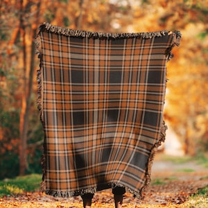 Puede incluir: Una manta de tiro de cuadros marrones y beige con flecos en los bordes. La manta está drapeada sobre una persona de pie en un entorno boscoso.