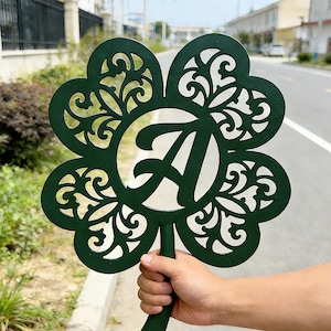 May include: A dark green metal garden stake shaped like a four-leaf clover with the letter "A" in the center. The clover has an ornate, openwork design. The stake is held up in front of a blurred outdoor background.