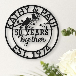 May include: Black metal wall art with the names "Kathy & Paul" and the words "50 Years Together EST. 1974". The design includes two birds perched on a branch within a circular frame.